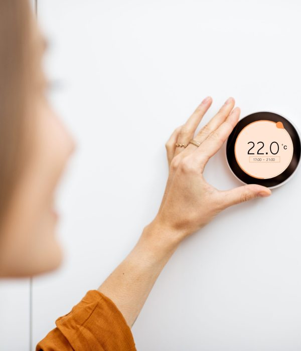 Young woman regulating heating temperature with a modern wireless thermostat installed on the white wall at home. Smart home heating regulation concept